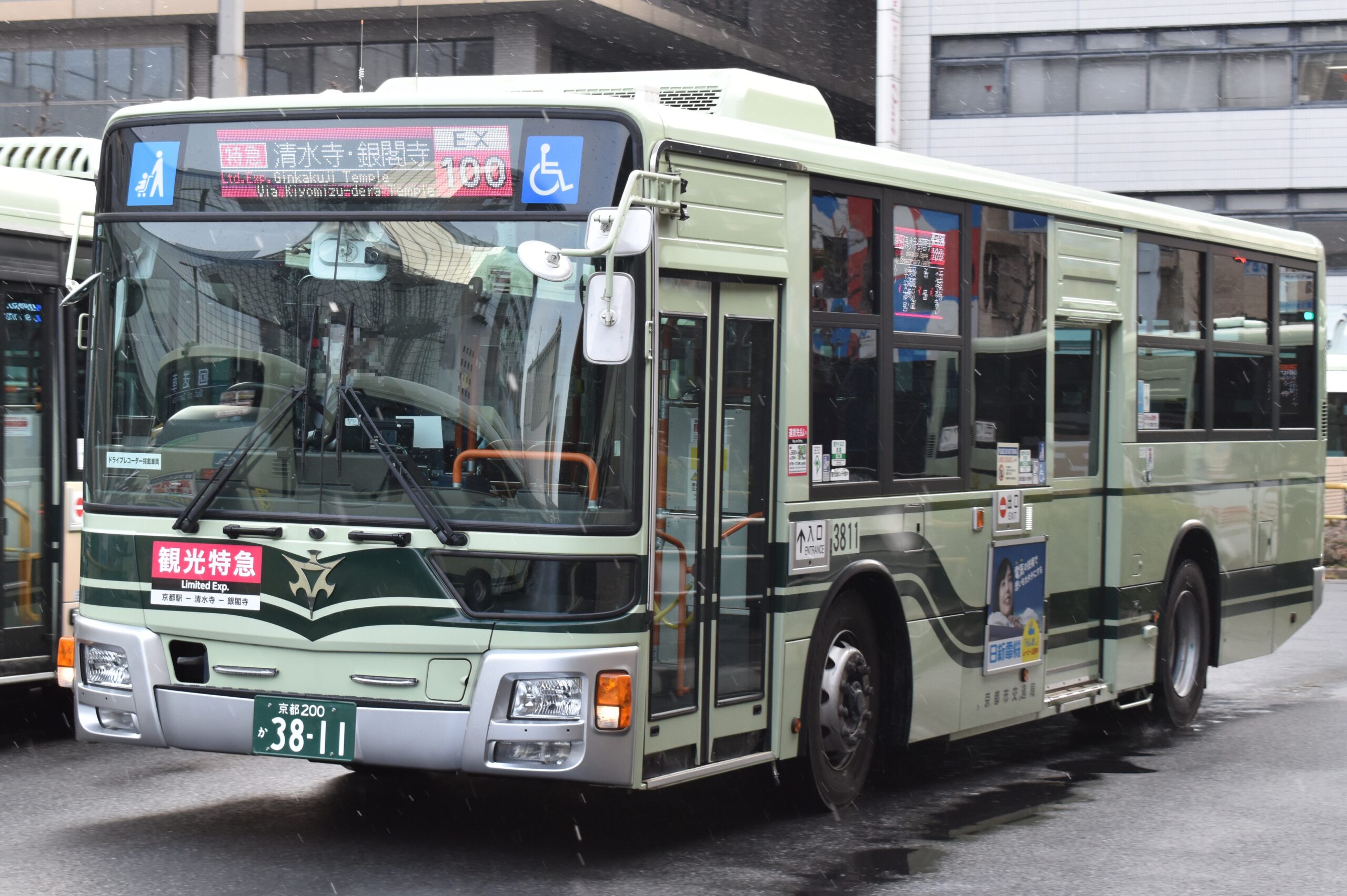 Bus express touristique EX100 du réseau municipal de Kyoto, vert et crème, affichant les destinations Kiyomizu-dera et Ginkaku-ji, à l'arrêt devant la gare de Kyoto sous la pluie.